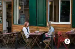 Tu ou vous ? Comment choisir le bon registre en français Deux femmes discutent à une table en terrasse d'un café en bois, entourées de chaises pliantes et d'une bouteille de vin avec verres posés sur la table.