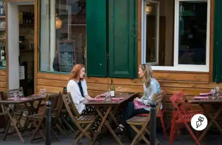 Tu ou vous ? Comment choisir le bon registre en français Deux femmes discutent à une table en terrasse d'un café en bois, entourées de chaises pliantes et d'une bouteille de vin avec verres posés sur la table.